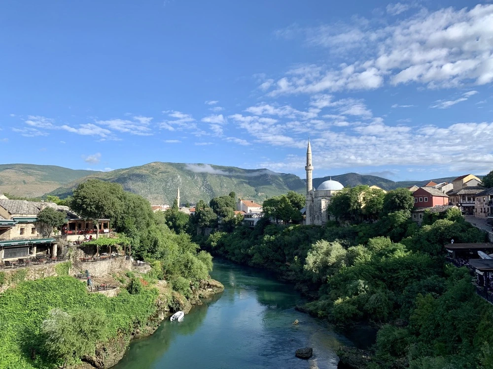 View from the Stari Most along the turquoise Neretva river in Mostar, with riverside terraces, a domed mosque and minaret, and the Herzegovina hills behind